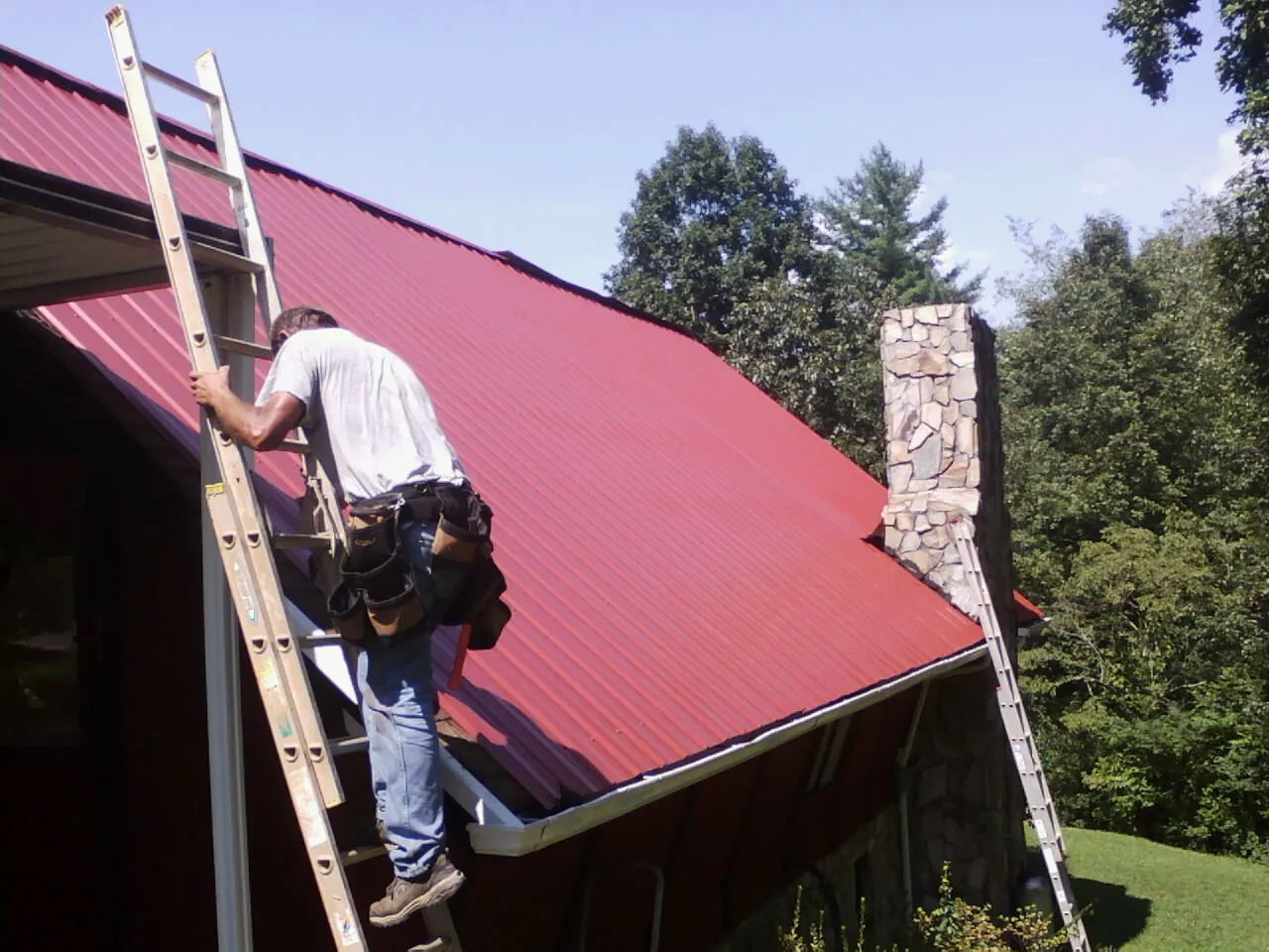 Roofer on a ladder beside a red metal roof and stone chimney — Soffit & Fascia Repair by Roofing Brothers in Silverthorne